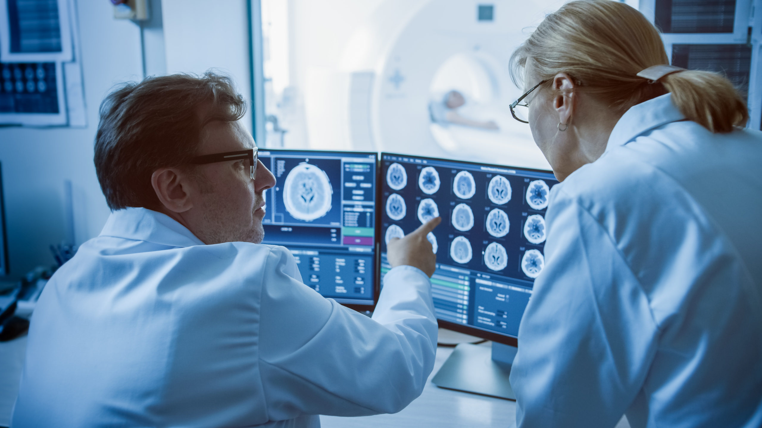 In Control Room Doctor and Radiologist Discuss Diagnosis while Watching Procedure and Monitors Showing Brain Scans Results, In the Background Patient Undergoes MRI or CT Scan Procedure.